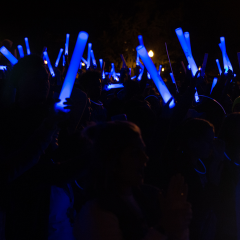 Attendees at the Blue Light Party at Indiana State University hold up glowing blue sticks in a darkened outdoor setting, creating a vibrant, energetic atmosphere.